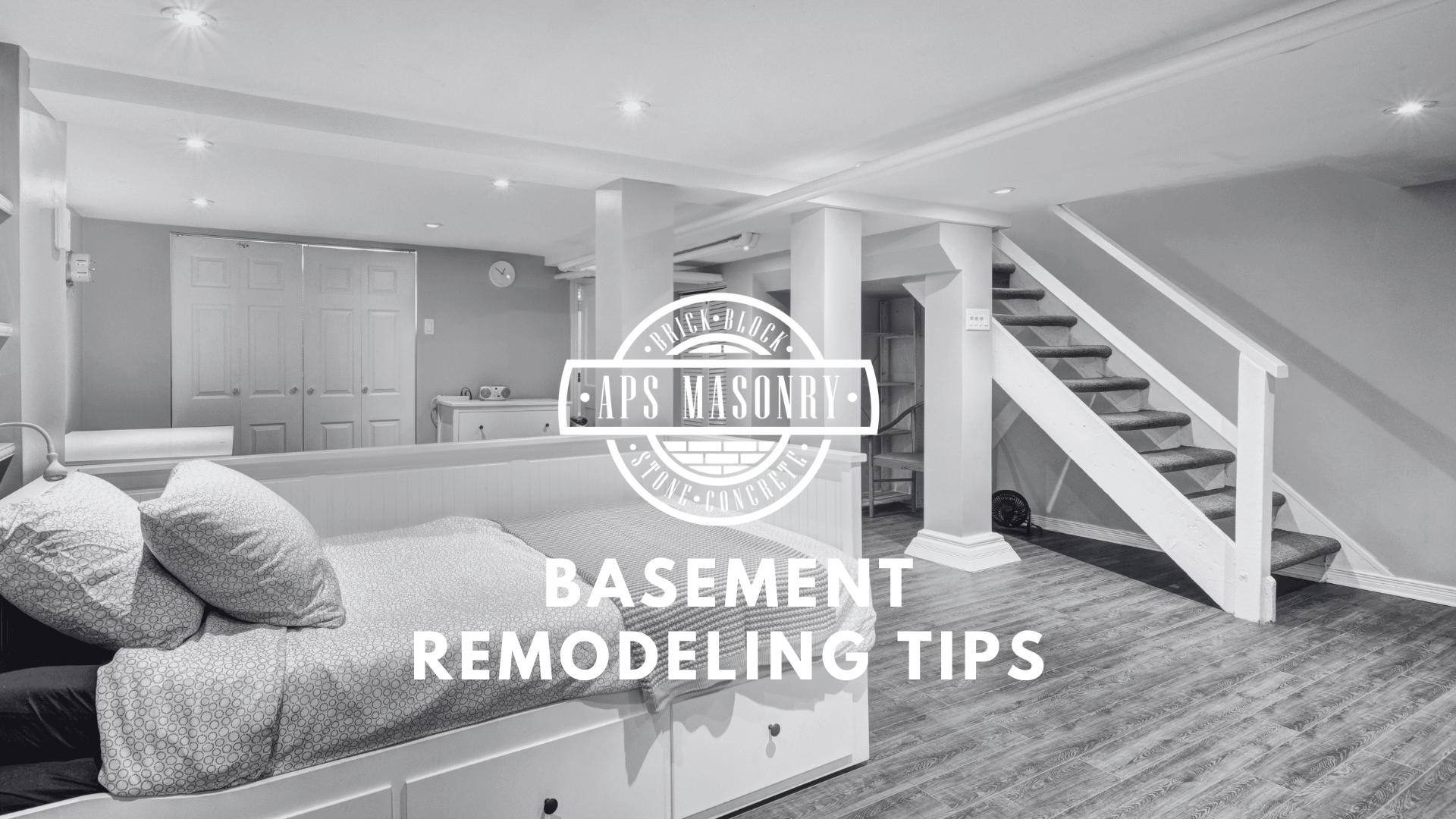 An image featuring a remodeled basement with a cozy nook with cushions, a built-in bench, and a neatly organized laundry area. In the foreground, there's a stylish wooden floor leading to a white staircase. The image is in black and white, giving it a modern and clean look. Overlaying the center is the logo of APS Masonry Contracting, and the text 