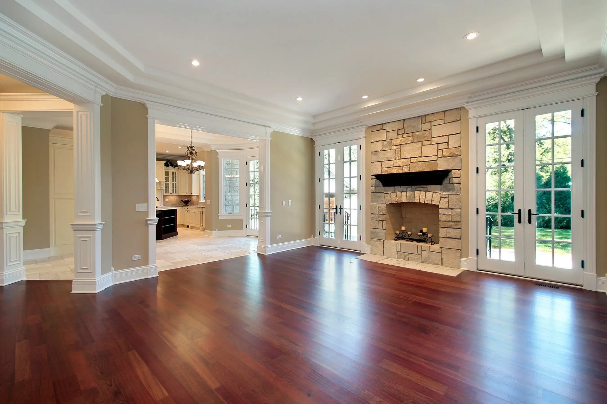 Spacious living room with hardwood floors and a stone fireplace, featuring large French doors that open to a bright outdoor view