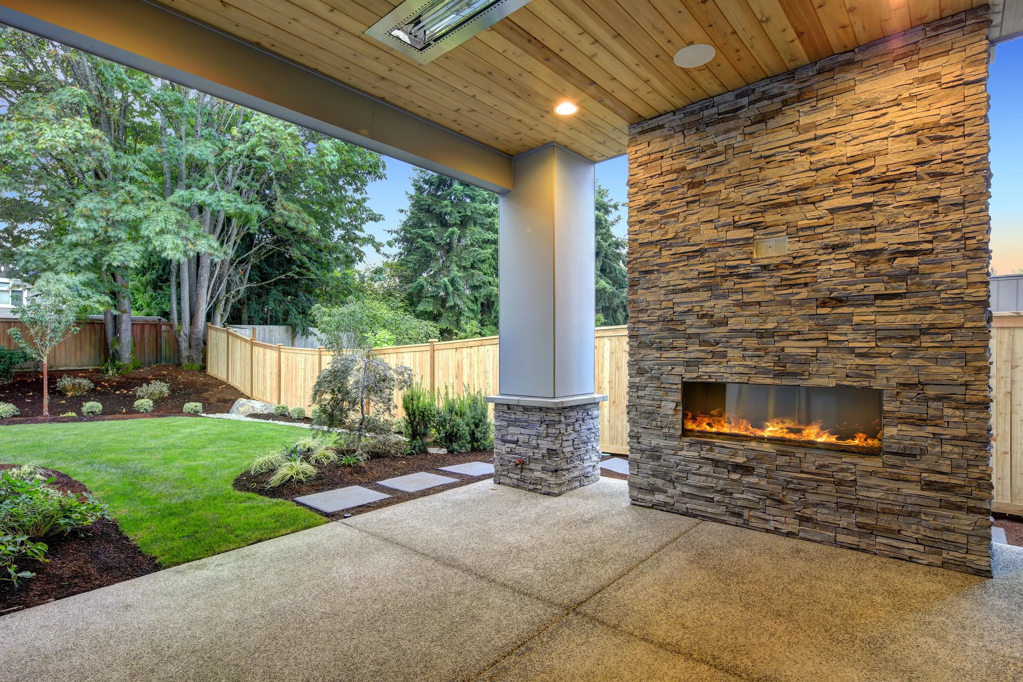 Modern covered patio with a stone-clad fireplace and a view of a landscaped backyard featuring a wooden fence and lush greenery