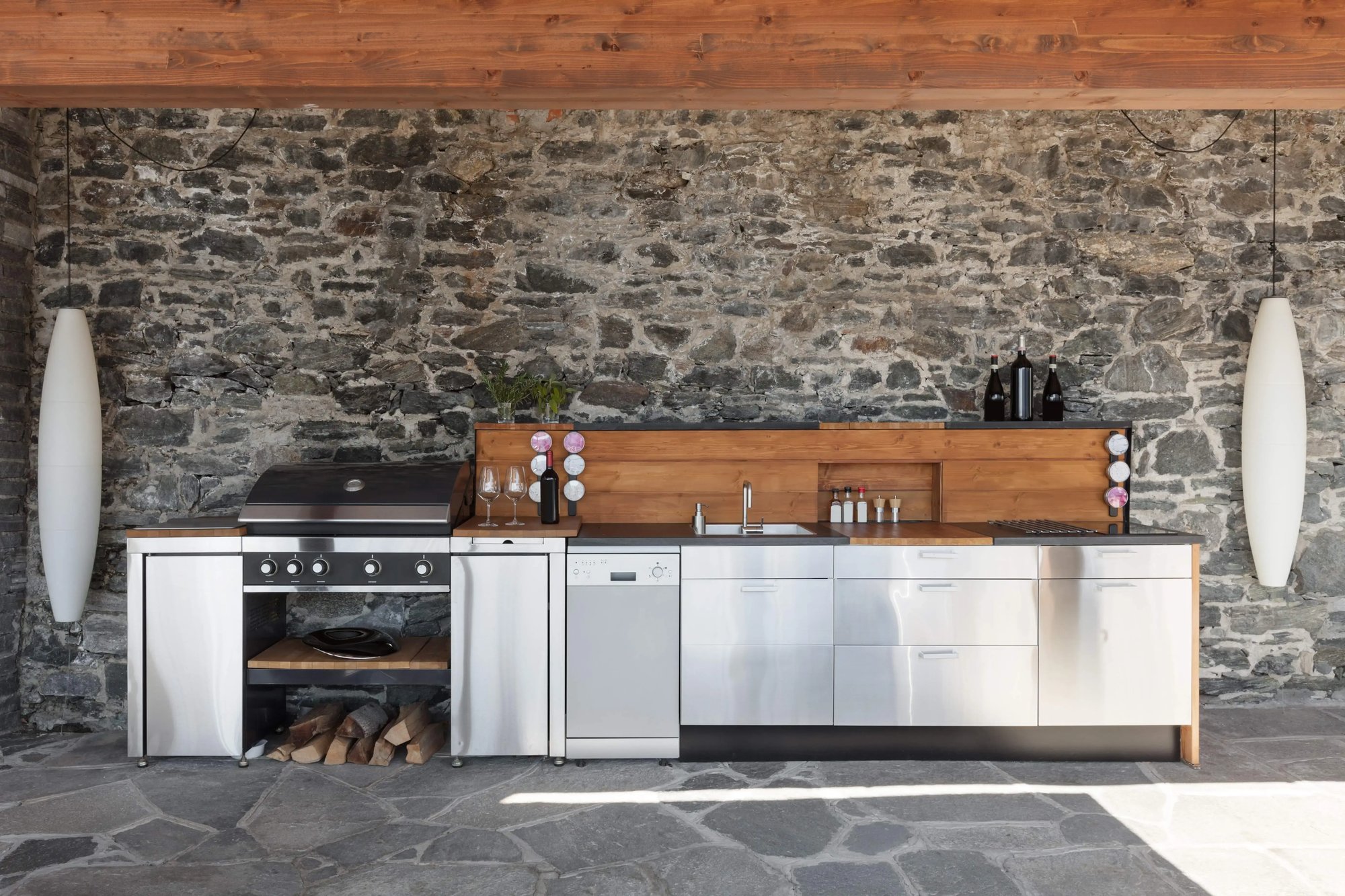 Modern outdoor kitchen with stainless steel appliances and wooden accents, set against a rustic stone wall