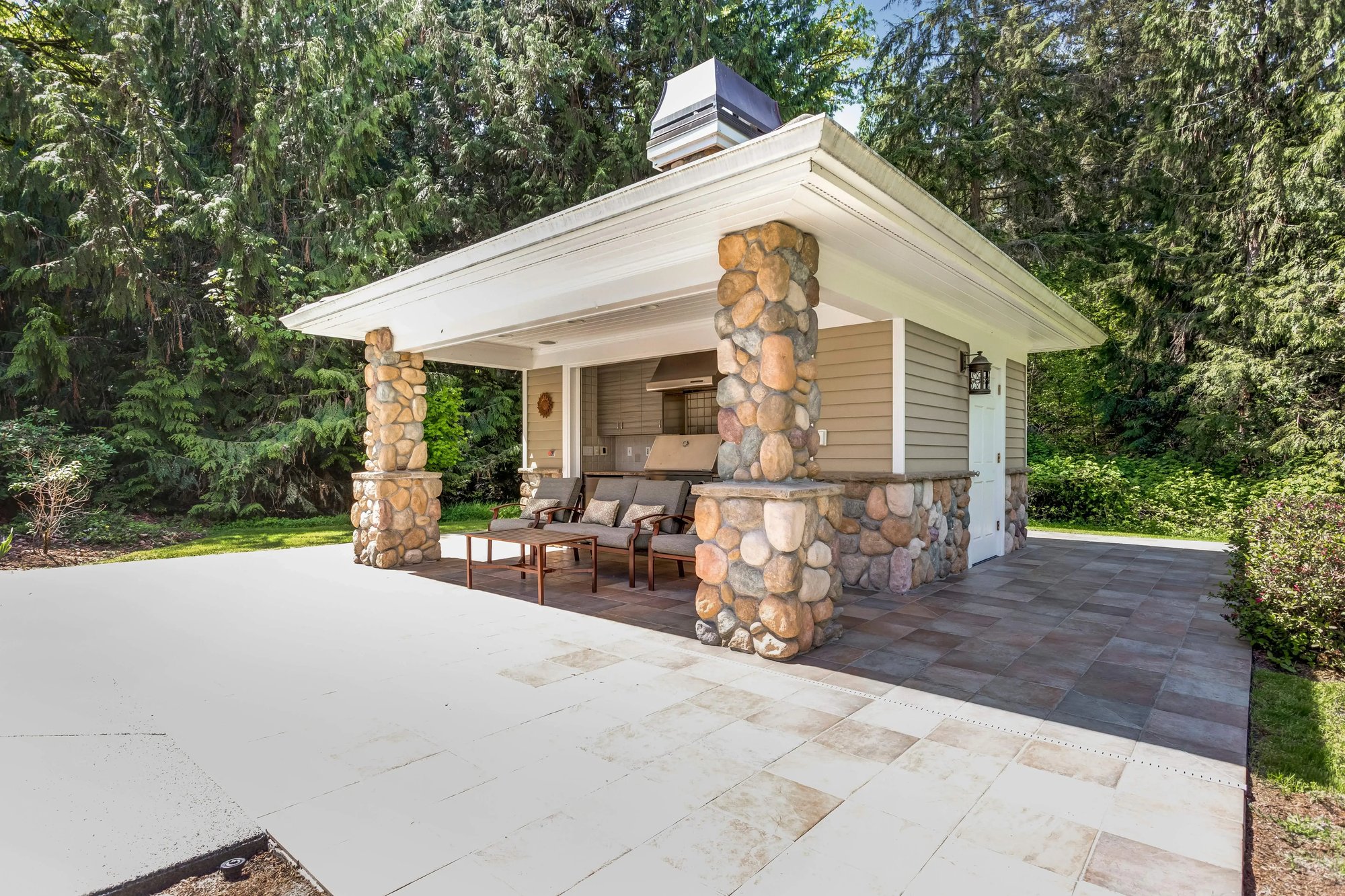 Outdoor patio with a covered seating area supported by stone columns, surrounded by a forested backdrop