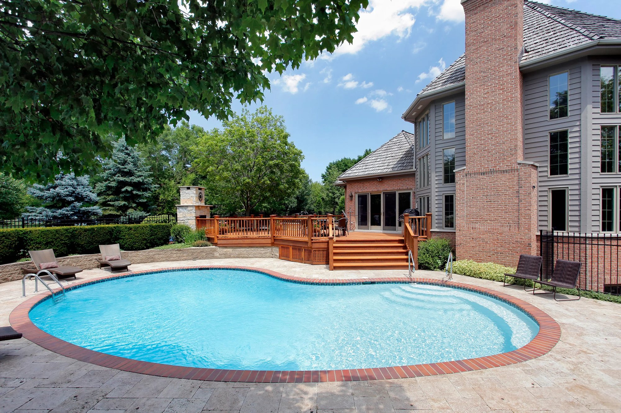 Newly renovated inground pool with brick border, wooden deck, and a large brick house in the background, surrounded by a beautifully landscaped yard