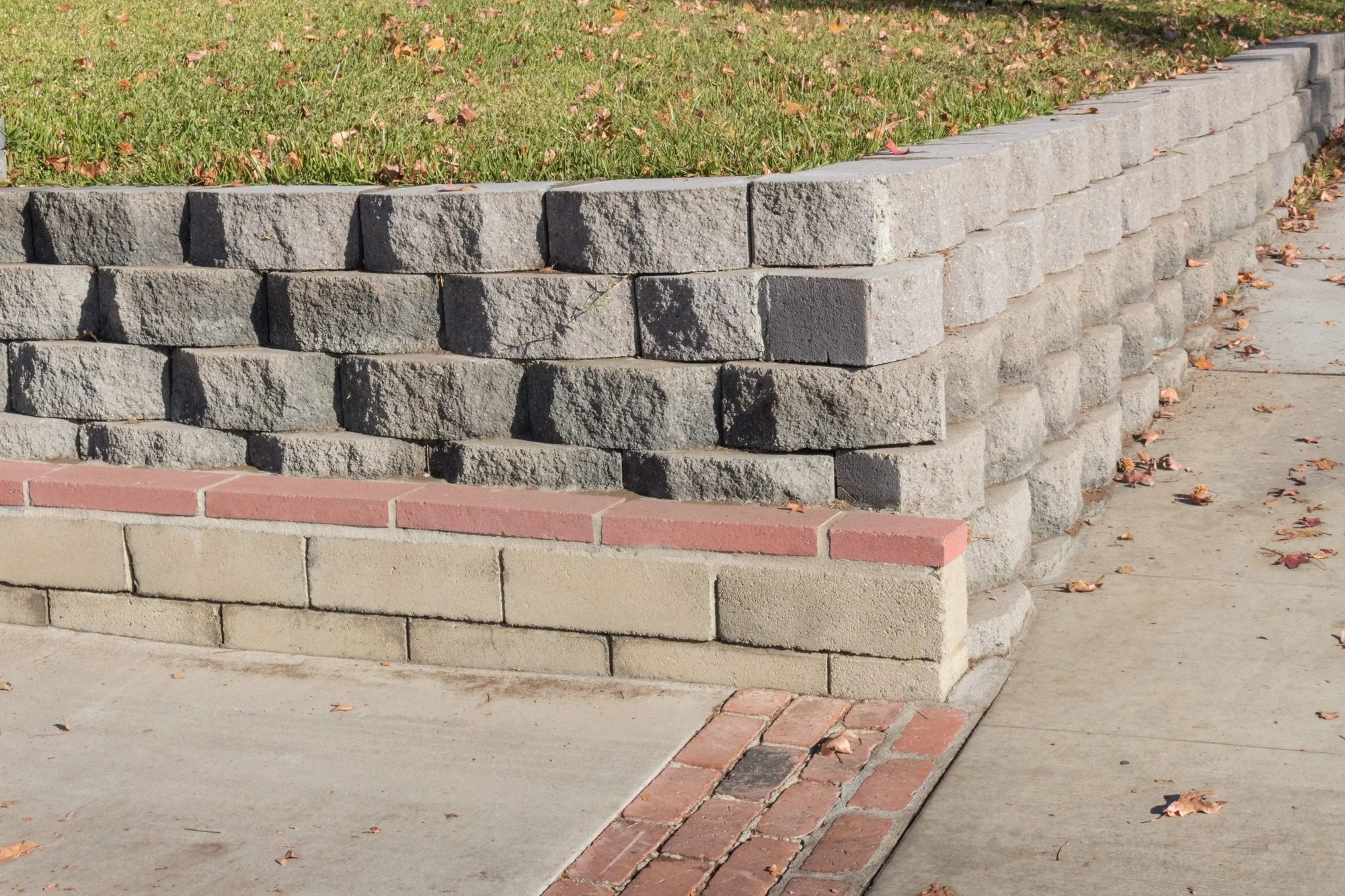 A sturdy retaining wall constructed with interlocking concrete blocks, featuring a decorative brick accent and sidewalk edge, perfect for landscape elevation control